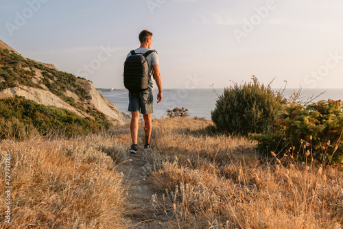 Hiker man with backpack hiking on the ocean coastline with mountains and evening sunlight.