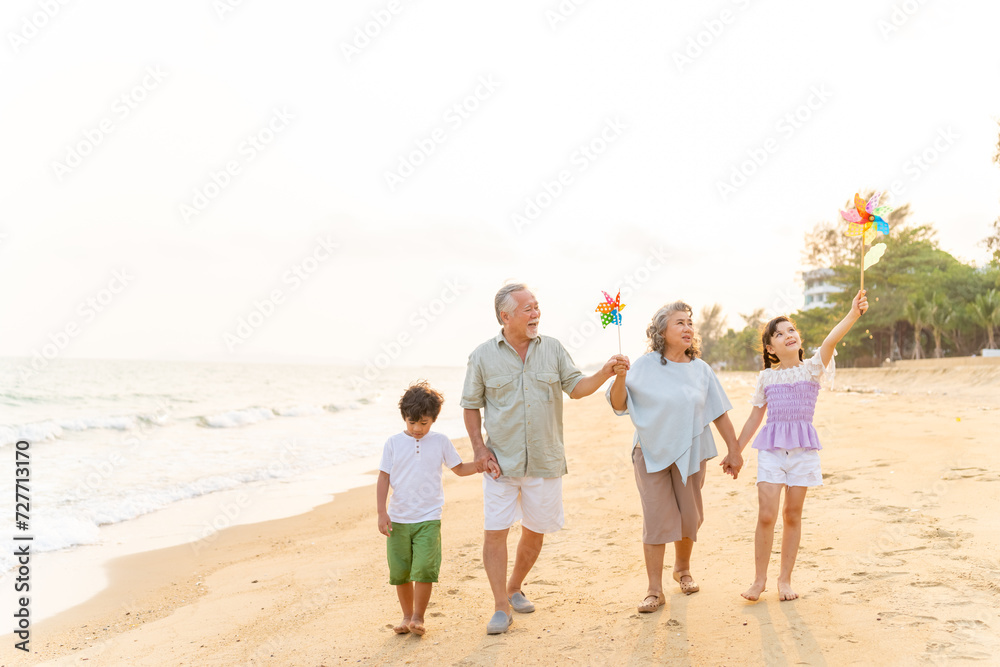 Happy Asian family travel ocean on summer holiday vacation. Grandparents and grandchildren boy and girl enjoy and fun outdoor lifestyle walking and playing together at tropical island beach at sunset.