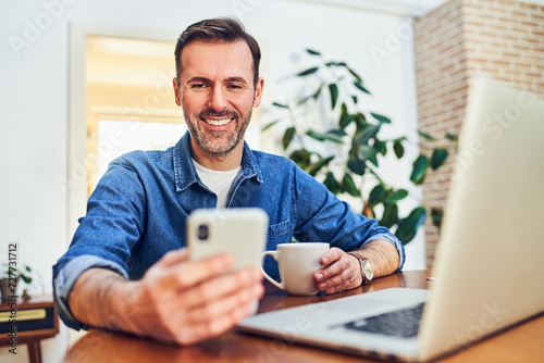 Cheerful man checking his mobile phone while working on laptop at home