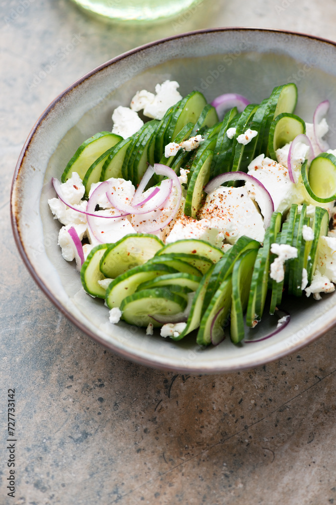 Salad with fresh cucumber, feta cheese and red onion in a grey bowl, vertical shot on a light-brown granite background, middle close-up