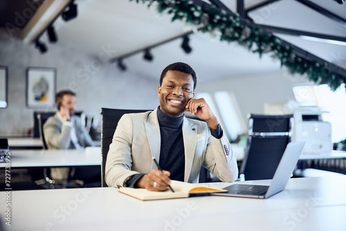A black businessman smiling for the camera, making some notes while his male colleagues are making a phone call in the background.