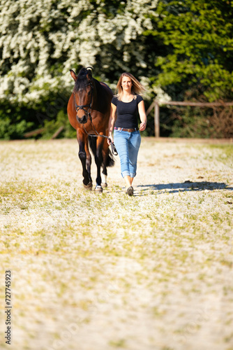 Fototapeta Naklejka Na Ścianę i Meble -  Young woman with a black shirt and short highlighted hair stands with her horse on a sunny riding arena.