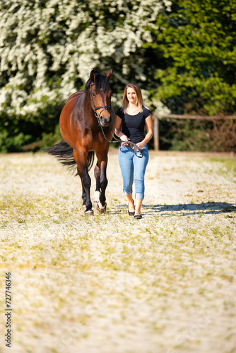 Young woman with a black shirt and short highlighted hair stands with her horse on a sunny riding arena. © RD-Fotografie