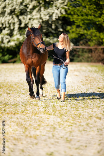 Young woman with a black shirt and short highlighted hair stands with her horse on a sunny riding arena.