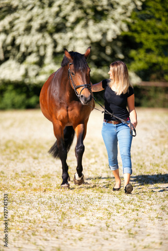Young woman with a black shirt and short highlighted hair stands with her horse on a sunny riding arena.