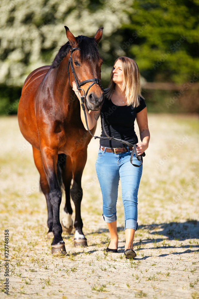 Obraz premium Young woman with a black shirt and short highlighted hair stands with her horse on a sunny riding arena.