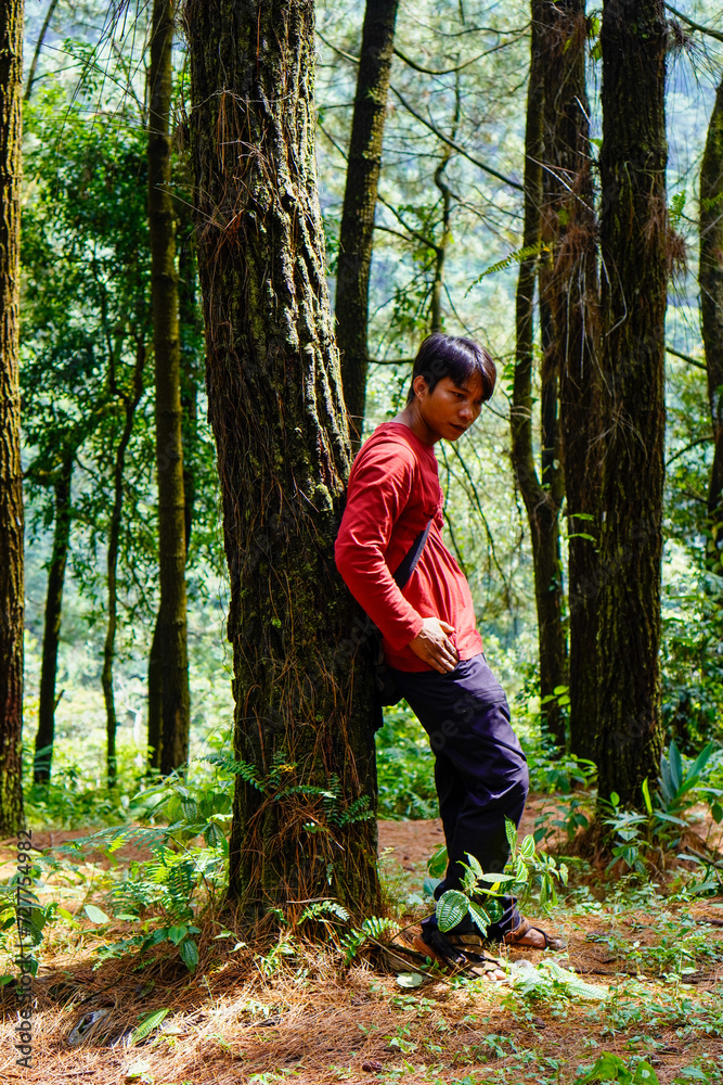 Javanese man in red is leaning against a tree in the forest. Javanese man in red is leaning against a tree in the forest.