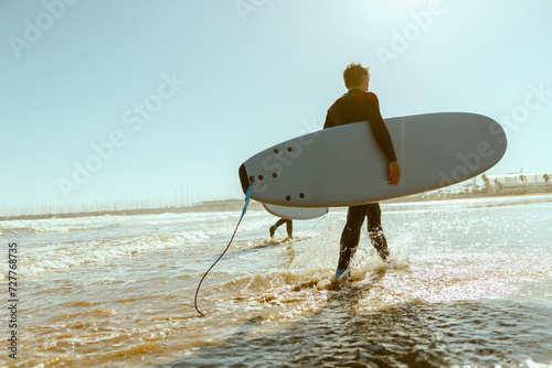 Back view of male surfer in wetsuit with his surfboard entering the sea. Surfing on ocean