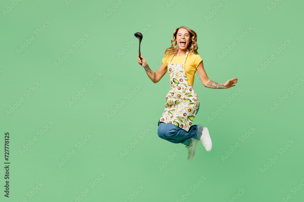 Full body smiling young housewife housekeeper chef cook baker woman wear apron yellow t-shirt jump high with ladle dipper look camera isolated on plain green background studio. Cooking food concept.