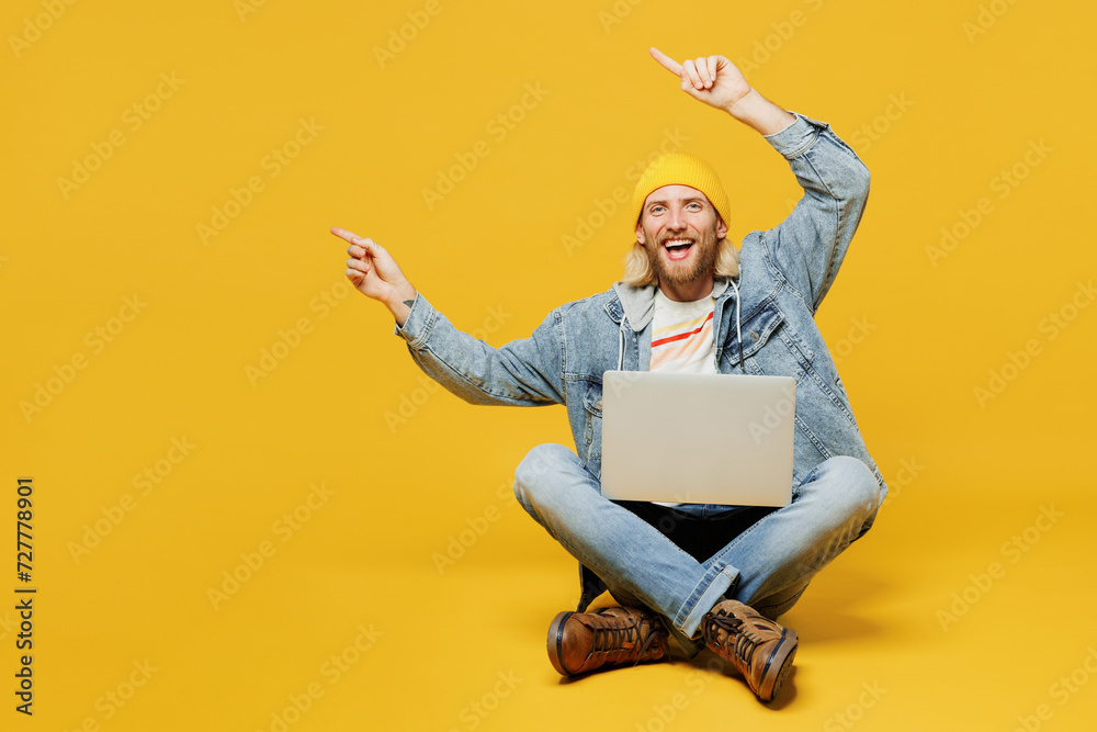 Full body young IT man wears denim shirt hoody beanie hat casual clothes sitting hold use work on laptop pc computer point aside isolated on plain yellow background studio portrait. Lifestyle concept. Full body young IT man wears denim shirt hoody beanie hat casual clothes sitting hold use work on laptop pc computer point aside isolated on plain yellow background studio portrait. Lifestyle concept.