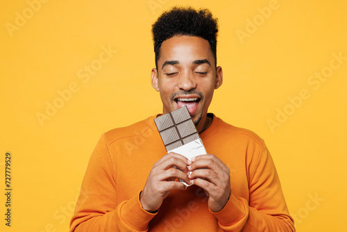 Young fun man wear orange sweatshirt casual clothes hold eat biting bar of chocolate close eyes isolated on plain yellow background studio. Proper nutrition healthy fast food unhealthy choice concept.