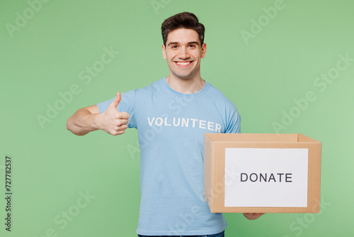 Young man wears blue t-shirt white title volunteer hold cardboard donation box show thumb up isolated on plain pastel green background. Voluntary free team work assistance help charity grace concept. photo