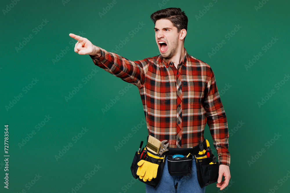 Young sad mad angry employee laborer handyman man wear red shirt point index finger aside scream isolated on plain green background. Instruments accessories, renovation apartment. Repair home concept. Young sad mad angry employee laborer handyman man wear red shirt point index finger aside scream isolated on plain green background. Instruments accessories, renovation apartment. Repair home concept.