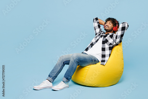 Full body young Indian man he wears shirt white t-shirt casual clothes sit in bag chair hold hands behind neck listen to music in headphones isolated on plain blue cyan background. Lifestyle concept.