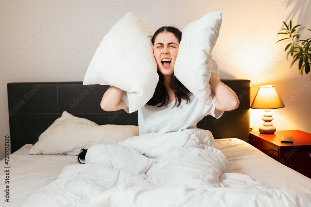 Portrait of annoyance Caucasian woman covers her ears with pillows and screams sitting in bed. Insomnia due to noisy neighbors Portrait of annoyance Caucasian woman covers her ears with pillows and screams sitting in bed. Insomnia due to noisy neighbors