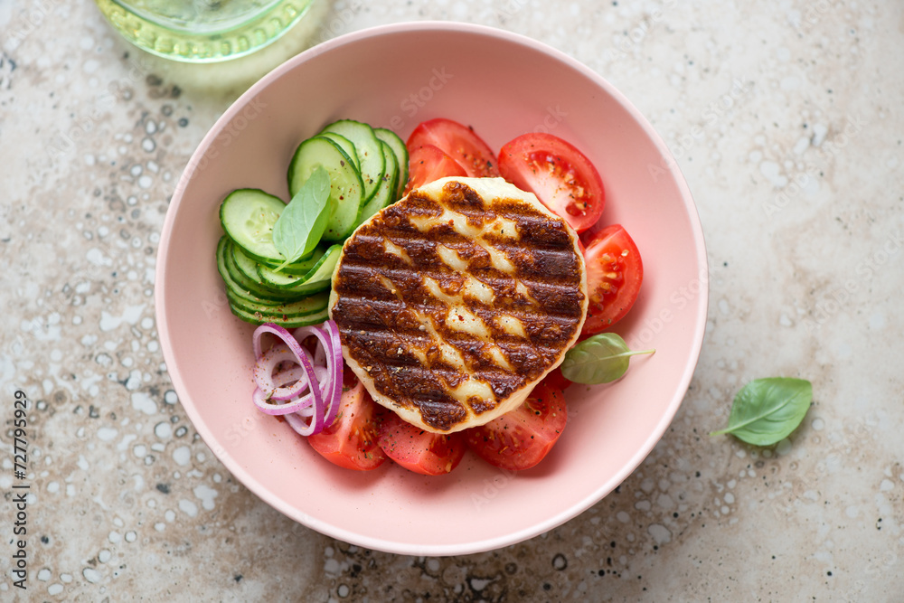 Roseate bowl with grilled haloumi cheese, tomatoes, cucumber and red onion, horizontal shot on a beige granite surface, high angle view