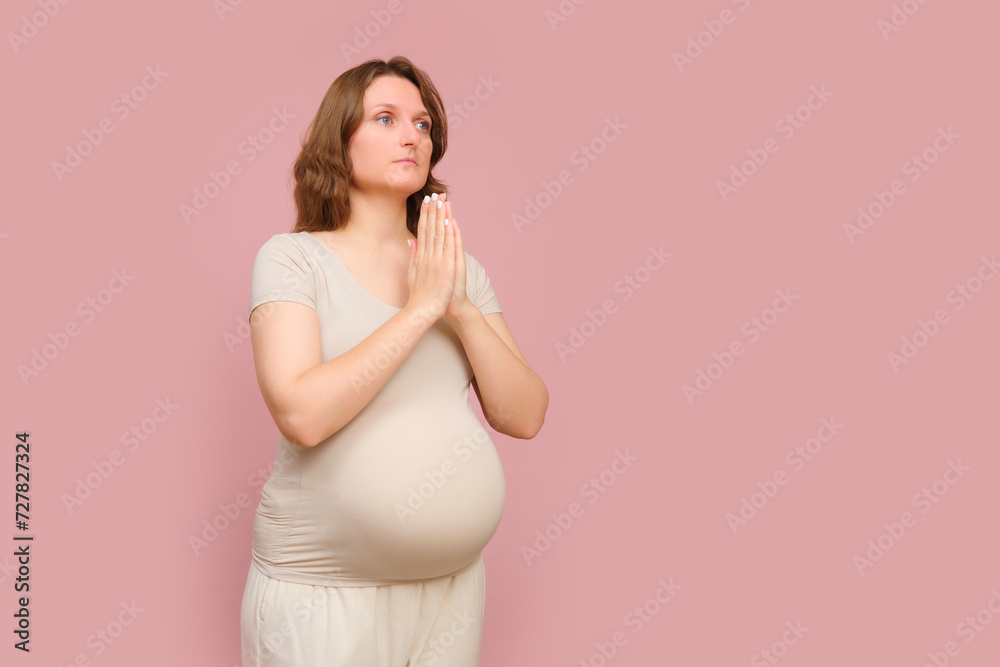 A pregnant woman prays on a studio pink background. Pregnancy in a woman with a belly and hands in a gesture of prayer, copy space A pregnant woman prays on a studio pink background. Pregnancy in a woman with a belly and hands in a gesture of prayer, copy space