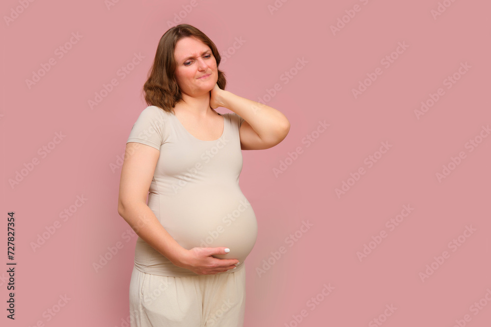 A pregnant woman holds onto a aching neck on a studio pink background. Pregnancy in a woman with a belly, copy space A pregnant woman holds onto a aching neck on a studio pink background. Pregnancy in a woman with a belly, copy space