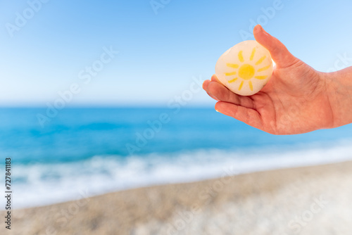 Child's hand holding a beach stone with a sun drawn on it, a defocused beach in the background.