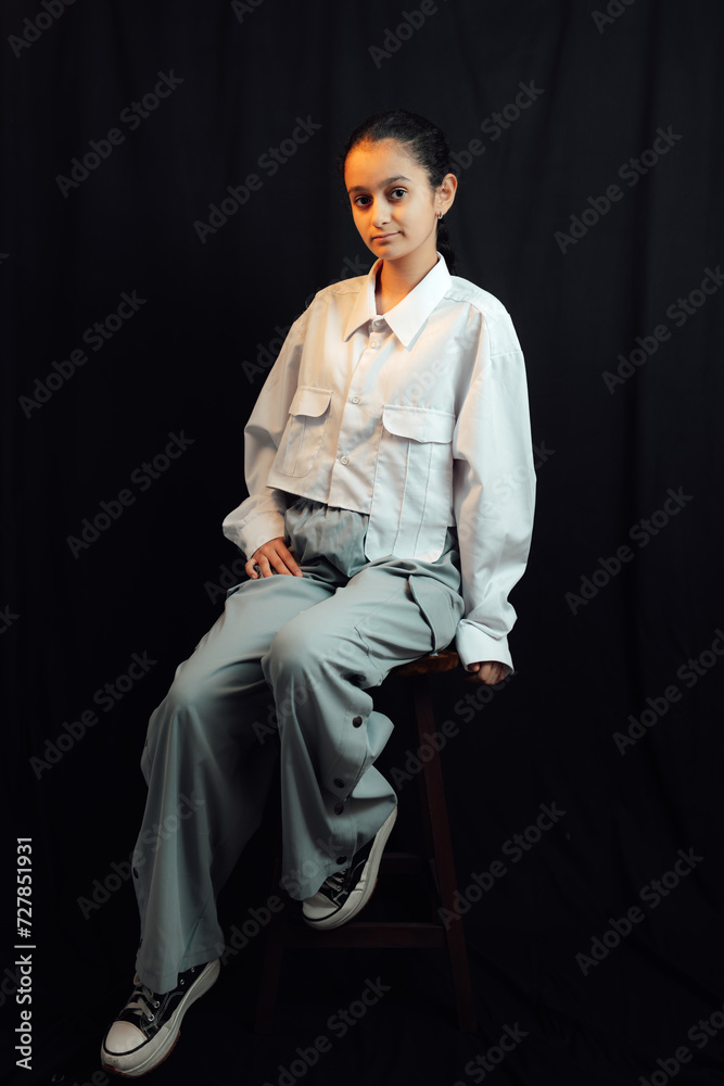 A studio shot of 13 years old fashionable girl sitting on the chair in front of black background and posing for camera