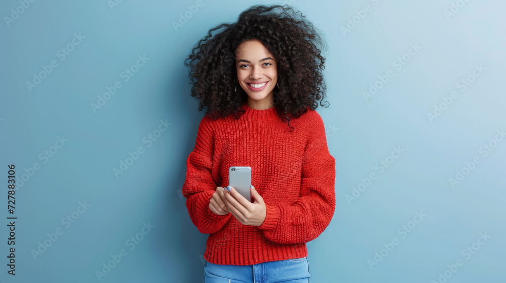 young woman with voluminous curly hair is smiling and holding a smartphone, wearing a red sweater, against a teal background young woman with voluminous curly hair is smiling and holding a smartphone, wearing a red sweater, against a teal background