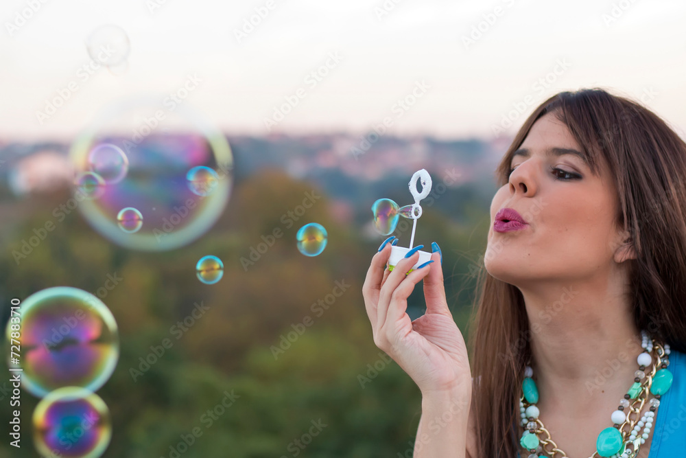Young attractive brunette with necklace making bubbles outdoors in the park. Back to the childhood