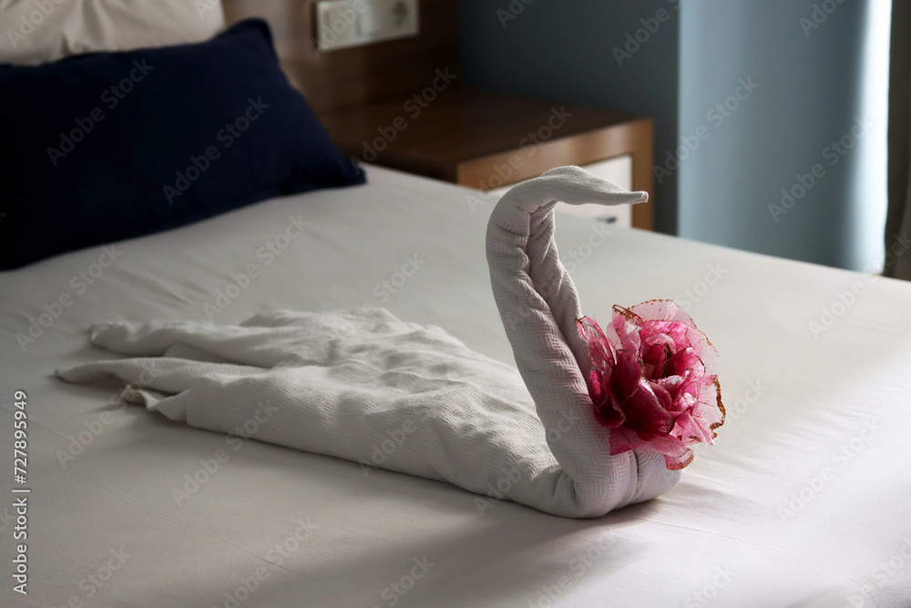 Elegant towel swan with floral decor on a hotel bed. Swan-shaped towel art with a pink flower on crisp linen. Creative towel origami swan with a flower on a white bedspread. Leisure and holidays