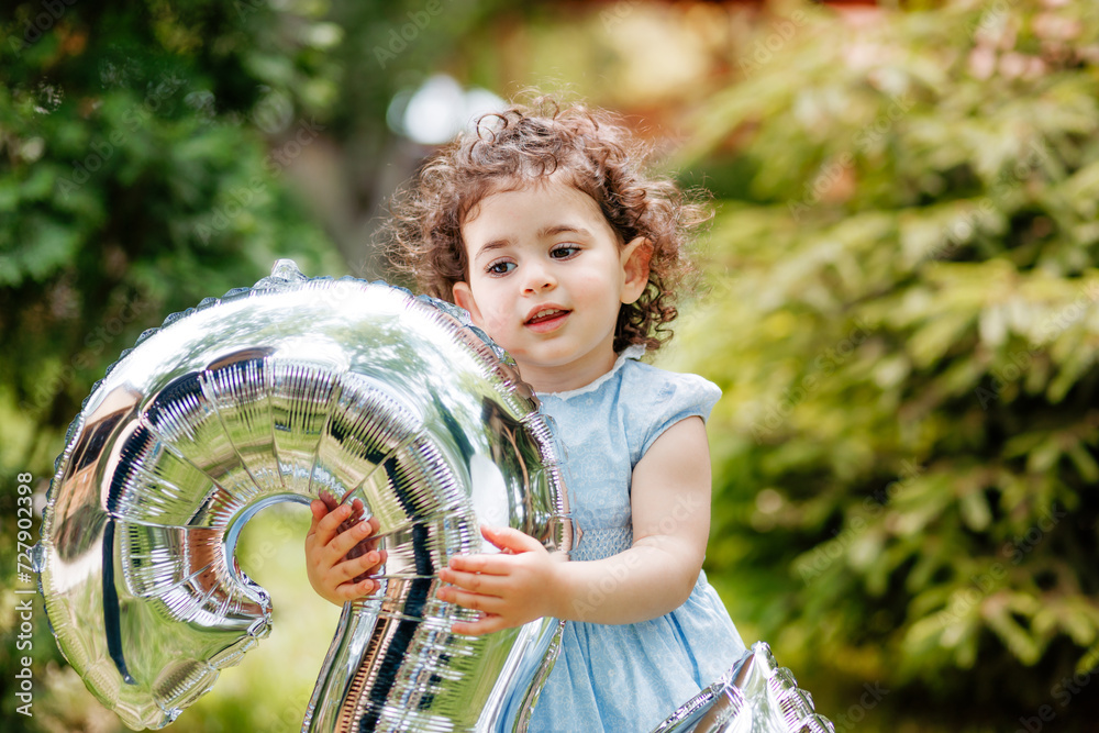 Cute two years old little girl with curly hair celebrating her birthday, holding balloon number