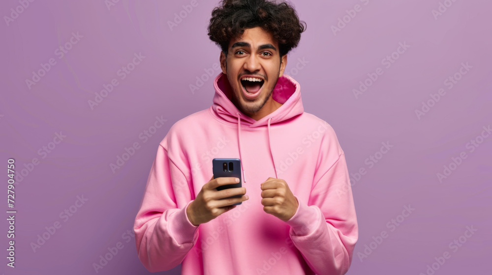 excited young man with curly hair in a pink hoodie, holding a smartphone with one hand and making a fist with the other excited young man with curly hair in a pink hoodie, holding a smartphone with one hand and making a fist with the other