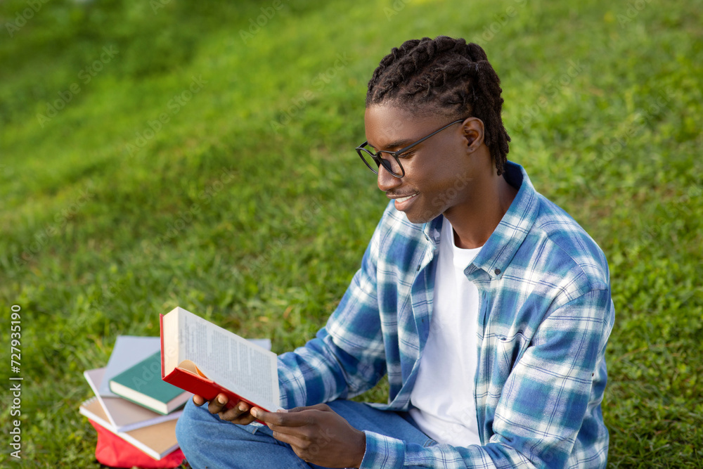 Fototapeta premium black man wearing glasses happily reads book relaxing in park
