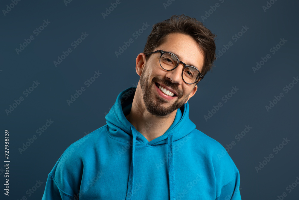 Smiling young man wearing glasses and bright hoodie standing against blue background Smiling young man wearing glasses and bright hoodie standing against blue background