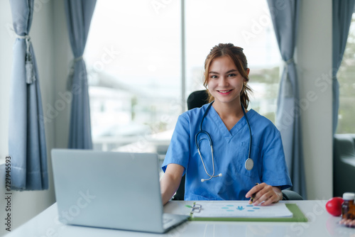 Friendly female nurse in blue scrubs seated at a desk, working on a laptop with medical charts and a stethoscope.. © amnaj