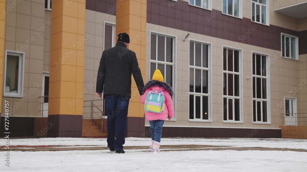 father takes child school holding his hand. happy family. snowy road school. father leads his little girl with school backpack through snow school. cold season. Dad holds hand child kid with backpack.
