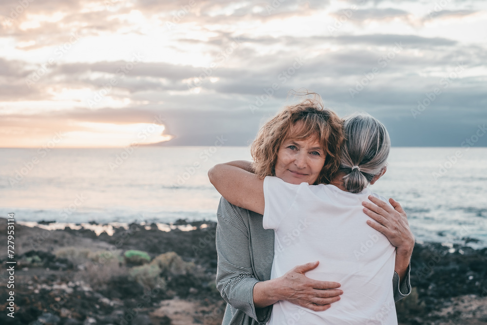 Fototapeta premium Romantic senior couple of women embrace each other on the seashore at sunset light, two embraced people stay together expressing love and tenderness