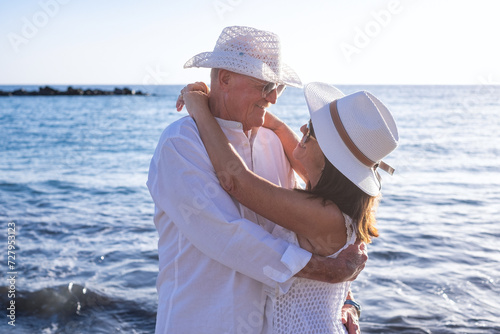 Cheerful bonding senior couple hugging on the seashore looking in the eyes enjoying vacation and sunset, two smiling people expressing love and tenderness