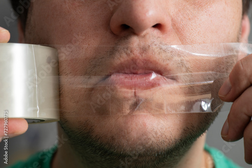 The man's mouth is sealed with duct tape. Transparent tape on the lips and in the hands of a man.
