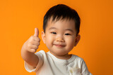 Cheerful Asian Toddler Giving Thumbs Up on Solid Orange Background