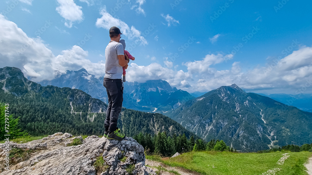Father holding baby with panoramic view from observation point of Monte Lussari, Camporosso, Friuli Venezia Giulia, Italy. Looking at mountain ridges of Julian Alps. Family trip on cloudy summer day