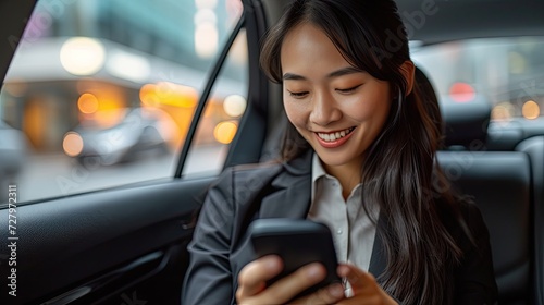 Asian woman sitting at the back of the car looking at her phone smiling, wearing black business suit with black hair. © MiguelAngel