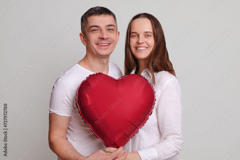 Smiling man and woman wearing white clothing holding heart shaped air balloon looking at camera smiling with happiness enjoying family anniversary isolated over gray background