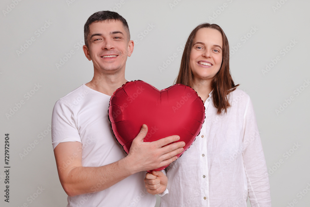 Pleased pretty couple looking at camera with smile satisfied man and woman wearing white clothing holding heart shaped air balloon isolated over gray background being in good mood