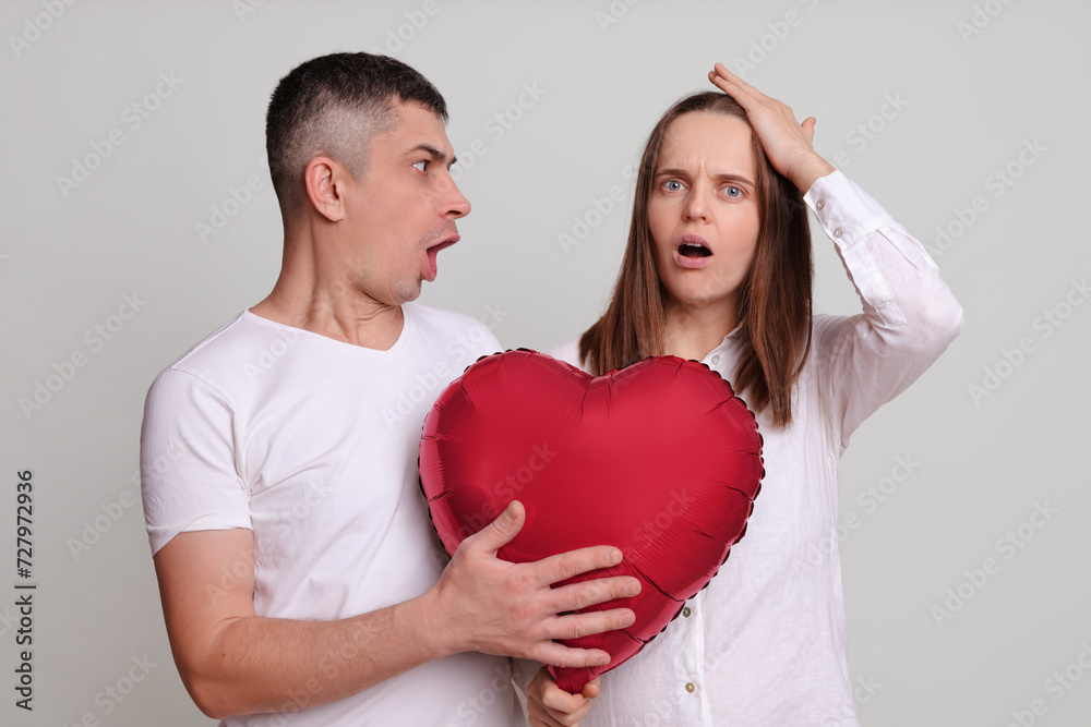 Astonished man and woman wearing white clothing holding heart shaped air balloon isolated over gray background female with facepalm gesture expressing negative feelings