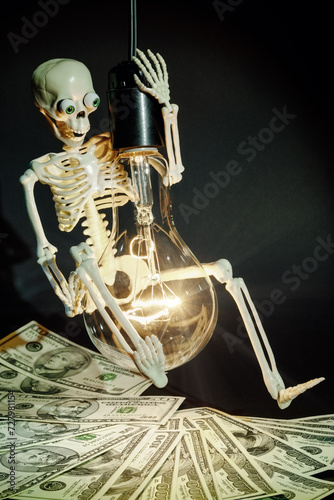A fun toy human skeleton swinging on a large incandescent light bulb above a pile of dollar bills. It symbolizes the upcoming high electricity prices and a more difficult life