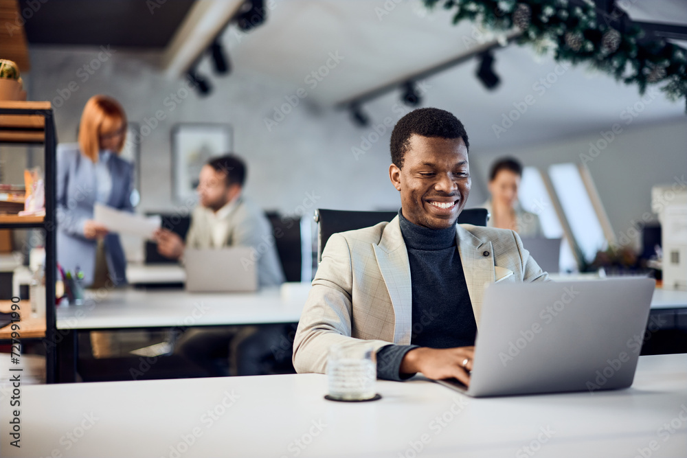 A smiling black businessman, working over the laptop, dressed in a suit, in a co-working space.