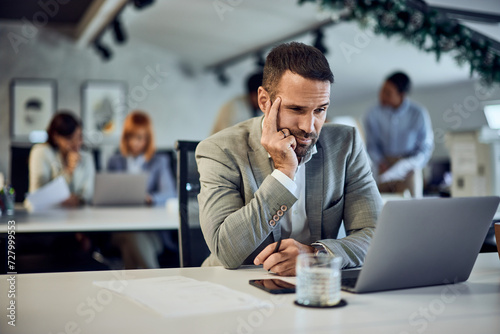 A focused businessman is working over the laptop, sitting in the co-working space, surrounded by his colleagues.