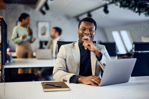 Portrait of a smiling black businessman, posing for the camera while sitting at the office and using a laptop.