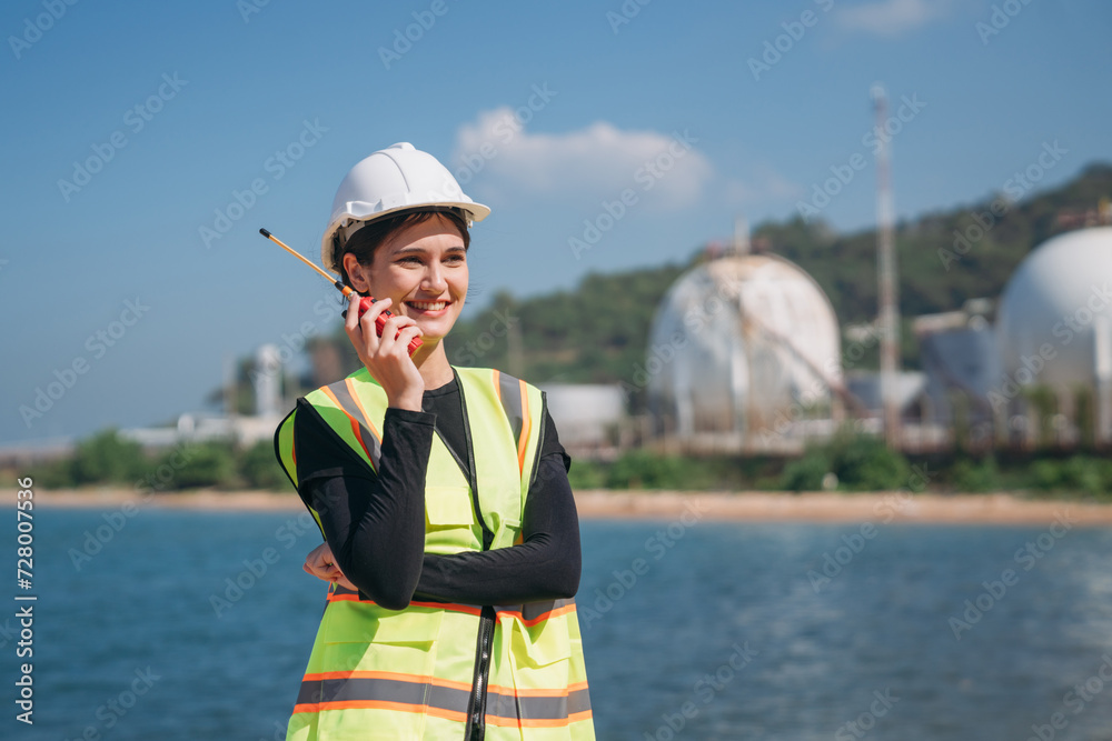 A young female engineer wearing a safety helmet and reflective vest holds a walkie-talkie at an industrial storage facility. A young female engineer wearing a safety helmet and reflective vest holds a walkie-talkie at an industrial storage facility.