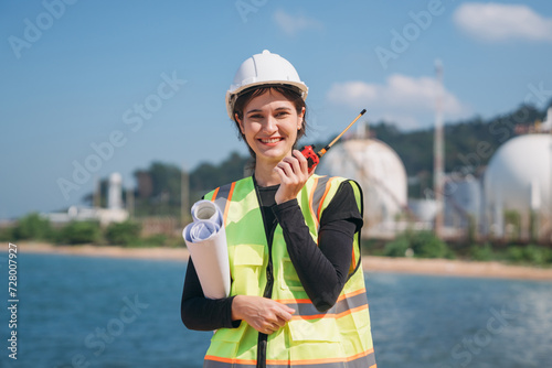 A young female engineer wearing a safety helmet and reflective vest holds a walkie-talkie at an industrial storage facility.