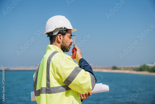Engineer wearing a safety helmet and reflective vest holds a walkie-talkie at an industrial storage facility.