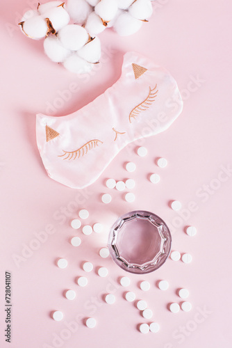 Good sleep concept, sleeping pills, glass of water and sleep mask on pink top and vertical view
